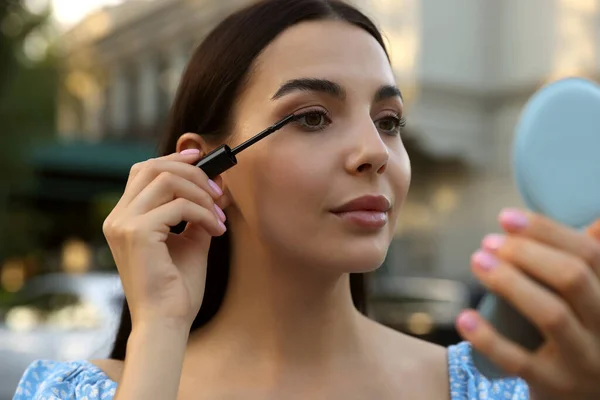 Beautiful woman with cosmetic pocket mirror applying mascara outdoors, closeup