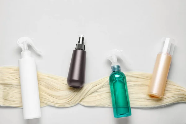 Spray bottles with thermal protection and lock of blonde hair on light background, flat lay