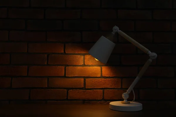 Stylish modern desk lamp on wooden table near brick wall at night, space for text