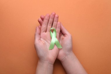 World Mental Health Day. Woman holding green ribbon on pale orange background, top view
