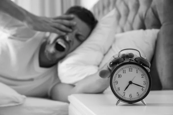 Sleepy man turning off alarm clock at home in morning, selective focus. Black and white photography