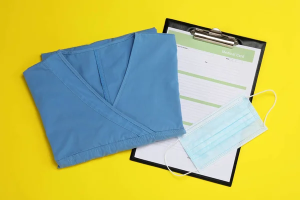 Medical uniform, face mask and clipboard on yellow background, flat lay