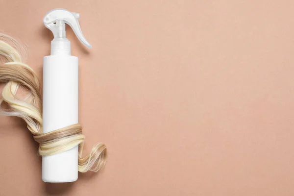 Spray bottle with thermal protection wrapped in lock of blonde hair on beige background, flat lay. Space for text