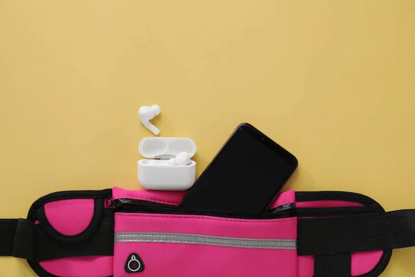 Stylish pink waist bag with smartphone and earphones on yellow background, flat lay. Space for text
