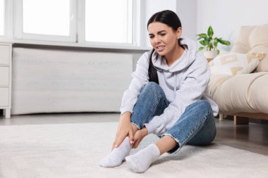 Woman suffering from ankle pain near sofa at home, space for text