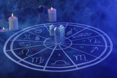 Zodyak işaretleri için doğal taşlar, astroloji çizelgesi ve koyu mavi masada yanan mumlar.
