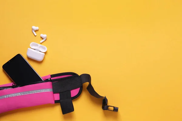 Stylish pink waist bag with smartphone and earphones on orange background, flat lay. Space for text