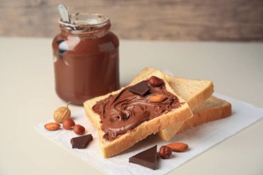 Tasty toast with chocolate paste and nuts near jar of cream on light table