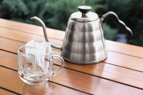 Glass cup with drip coffee bag and kettle on wooden table