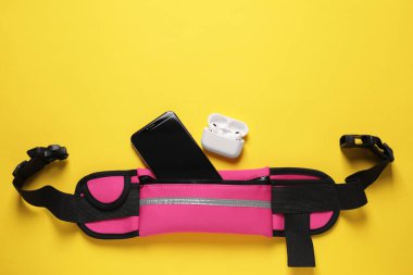 Stylish pink waist bag with smartphone and earphones on yellow background, flat lay. Space for text