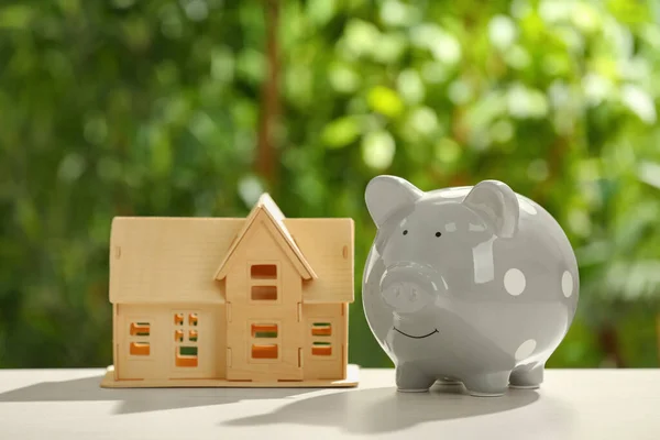 Piggy bank and house model on wooden table outdoors. Saving money concept