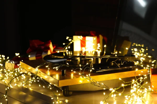 Turntable with vinyl record, fairy lights and Christmas gift boxes on grey table, closeup