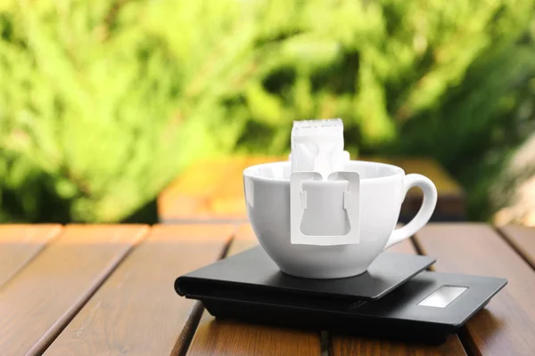 Cup with drip coffee bag and kitchen scales on wooden table outdoors