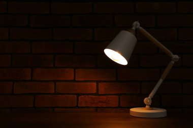 Stylish modern desk lamp on wooden table near brick wall at night, space for text