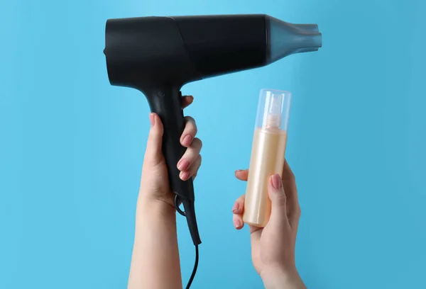Woman holding spray bottle with thermal protection and hairdryer on light blue background, closeup