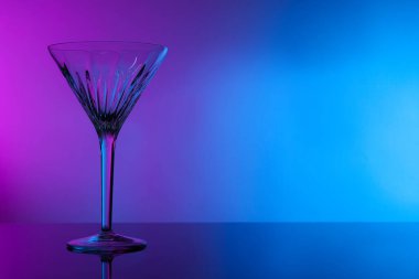 Empty clean martini glass on mirror table in neon lights, space for text