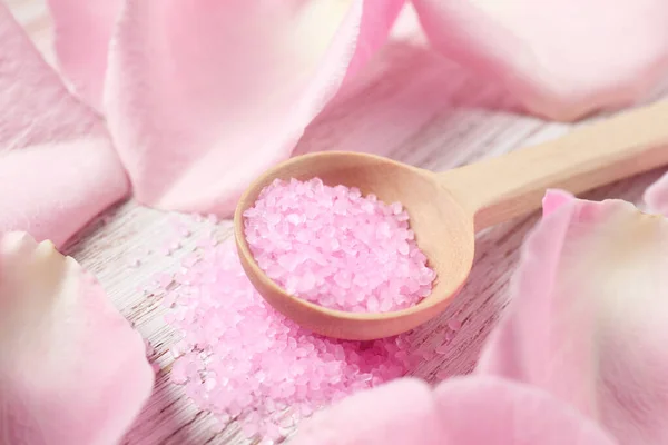 Spoon with pink sea salt and petals of roses on white wooden table, closeup