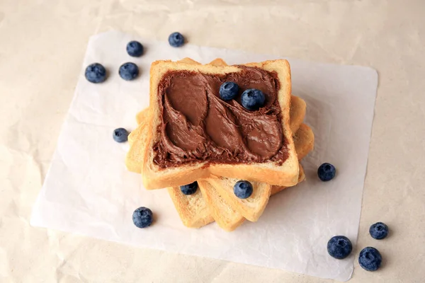 Tasty toast with chocolate paste and blueberries on parchment paper, above view