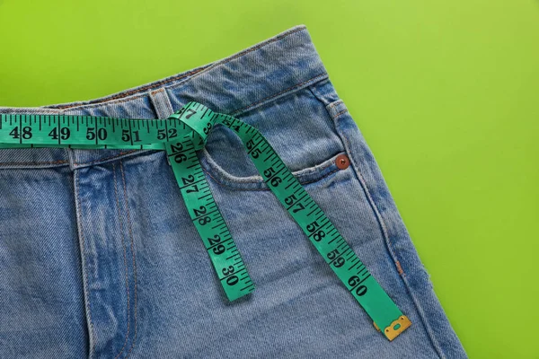 Jeans and measuring tape on light green background, top view. Weight loss concept