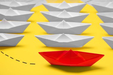 Red paper boat floating away from others on yellow background, closeup. Uniqueness concept