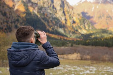 Boy looking through binoculars in beautiful mountains, back view. Space for text