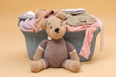 Laundry basket with baby clothes near soft toy on light brown background