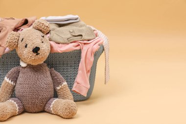 Laundry basket with baby clothes near soft toy on light brown background. Space for text