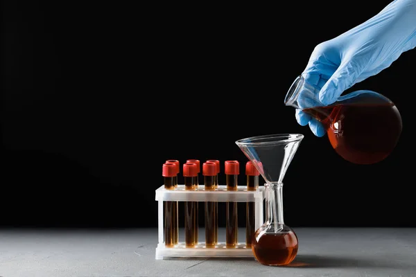 Scientist pouring brown liquid into flask at grey table against black background, closeup. Space for text