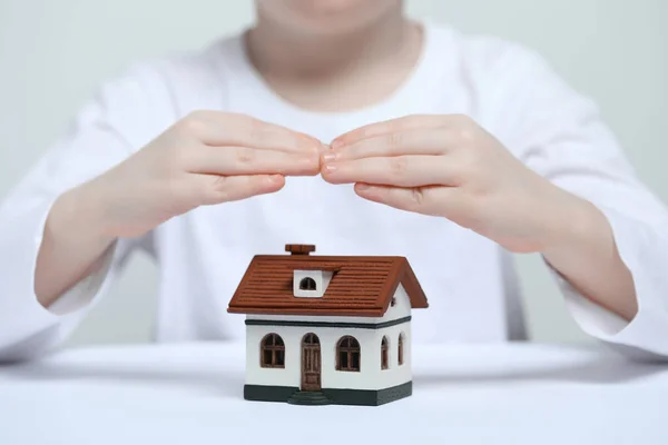 Home security concept. Little child covering house model at white table, closeup