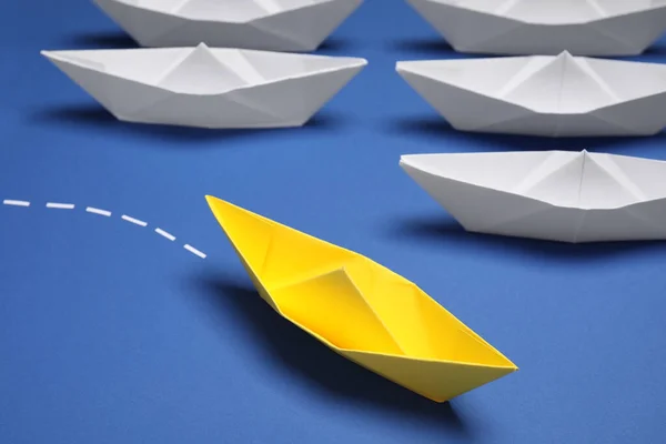 Yellow paper boat floating away from others on blue background, closeup. Uniqueness concept