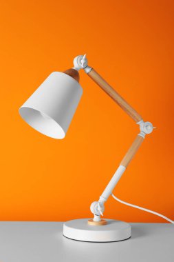 Stylish modern desk lamp on white table near orange wall