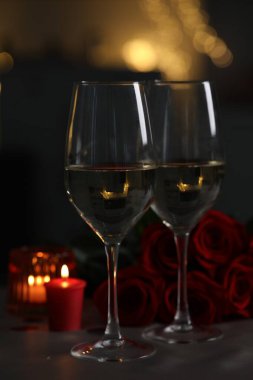 Glasses of white wine, burning candle and rose flowers on grey table against blurred lights. Romantic atmosphere