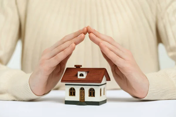 Home security concept. Woman covering house model at white table, closeup