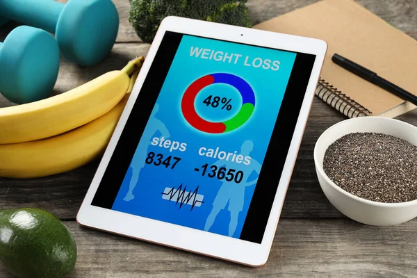 Tablet with weight loss calculator application, dumbbells and products on wooden table