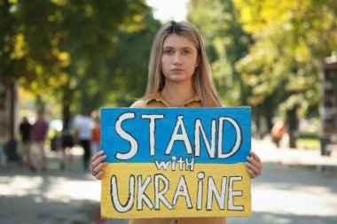 Sad woman holding poster in colors of national flag and words Stand with Ukraine outdoors