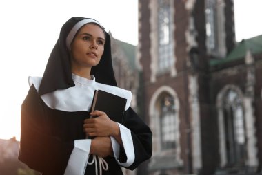 Young nun with Bible near cathedral outdoors on sunny day, space for text