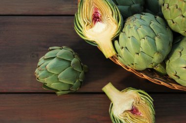 Wicker basket with fresh raw artichokes on wooden table, flat lay