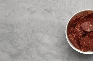 Delicious beef jerky in bowl on light gray table, top view. Space for text