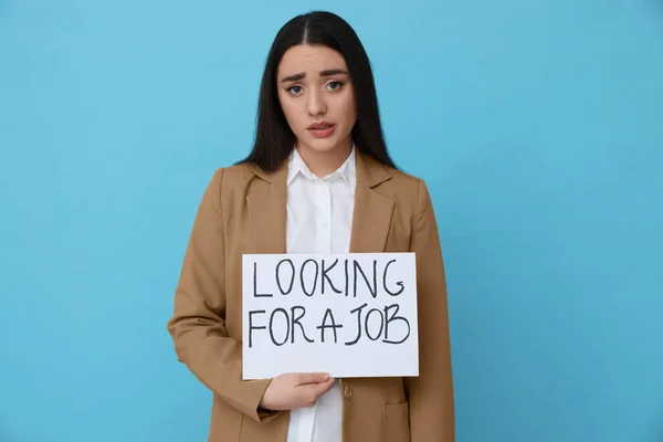 Young unemployed woman holding sign with phrase Looking For A Job on light blue background