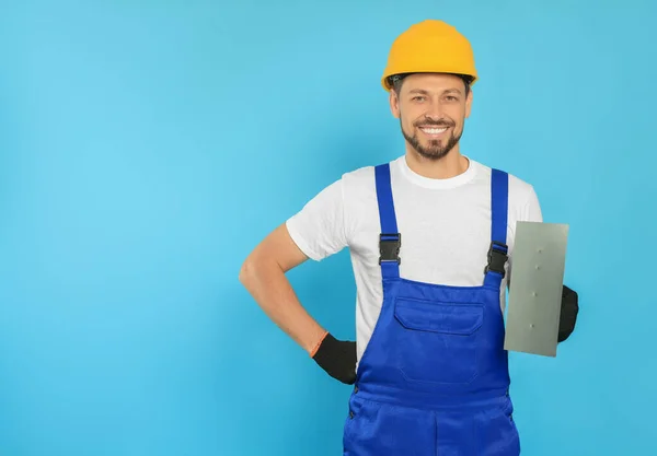 Professional worker in hard hat with putty knife on light blue background, space for text
