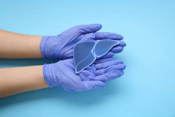 Doctor in gloves holding paper liver on light blue background, closeup and top view. Hepatitis treatment