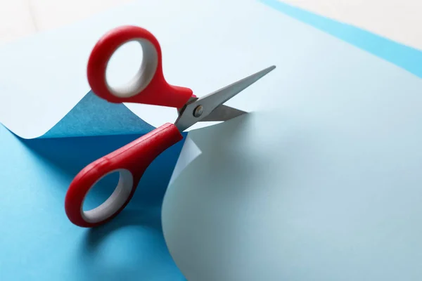 Red scissors cutting light blue paper on white background, closeup