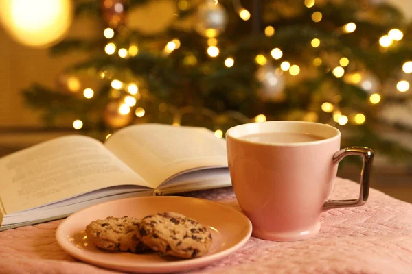 Cup of delicious cocoa, cookies and open book on pink fabric against blurred Christmas tree with lights