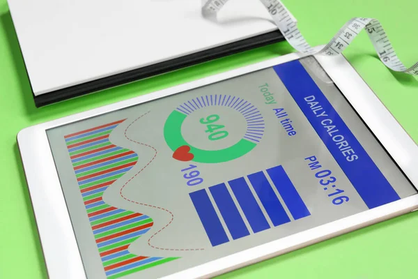 Tablet with weight loss calculator application and measuring tape on green background, closeup