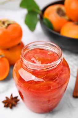 Jar of tasty persimmon jam and ingredients on white marble table, above view