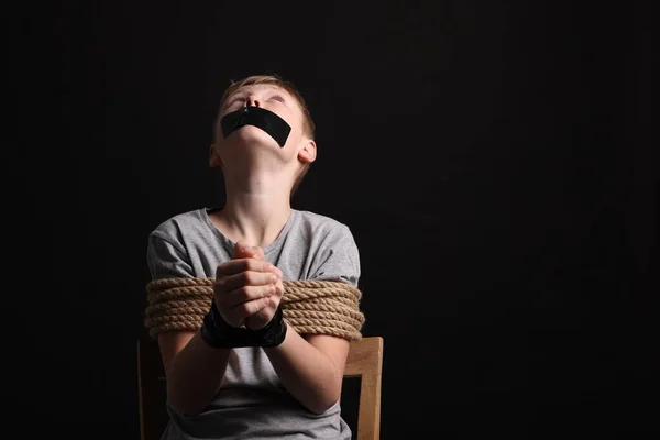 Little boy with taped mouth tied up and taken hostage against dark background. Space for text