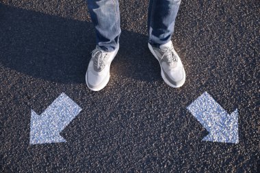 Choice of way. Man standing in front of drawn marks on road, closeup. White arrows pointing in different directions