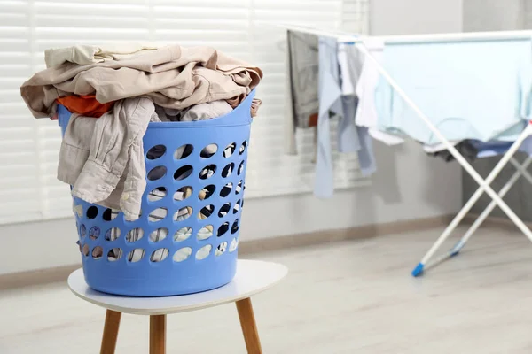 Plastic laundry basket overfilled with clothes on white stool indoors. Space for text