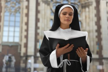 Young nun reading Bible near cathedral outdoors. Space for text