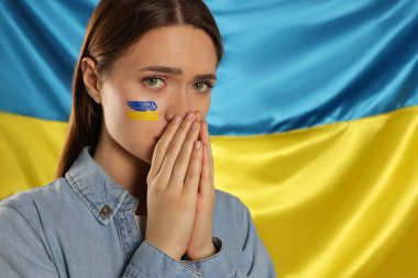 Sad young woman with clasped hands near Ukrainian flag. Space for text